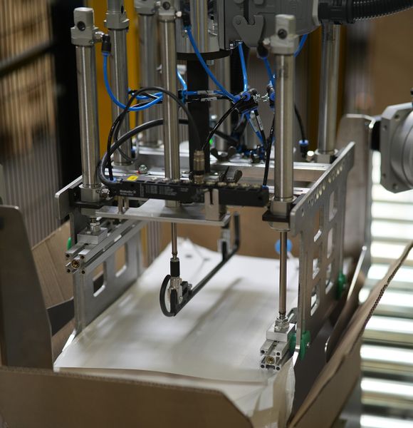 Bild e): Die Bilder a) bis e) zeigen den Transport eines Beutelstapels vom Ablageort über das Anbringen von Pappe und Stopfen sowie das Verschuppen des Bügels bis zur Ablage in den Karton. (Bild: Bihl+Wiedemann)