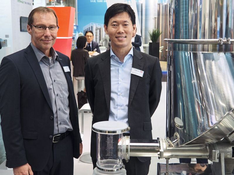 Guido Obler (links) und Yasushiro Kishihata, Brabender Kybota, neben dem NX-Feeder, der sich zur hochgenauen Dosierung für besonders schwerfließende Produkte eignet. (Bild: Mühlenkamp/SCHÜTTGUT)