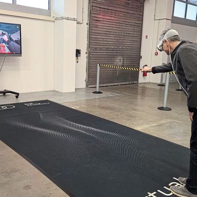 Mixed-Reality-Training bei Audi: Der Bildschirm im Hintergrund zeigt, was der Nutzer in der Brille sieht – gerade platziert er den Anschlussstutzen des Kühlmittel-Spülgerätes. Auf der schwarzen Fläche ist – nur für ihn sichtbar – das virtuelle Auto platziert. (Bild: Alina Seysen)