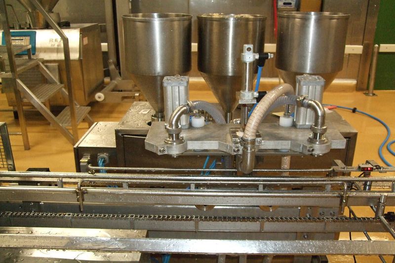 Die drei Fülltrichter sind mit jeweils einem cremeartigen Nahrungsmittelprodukt gefüllt. Sie sind auf dem Verfahrwagen angebracht, der sich synchron mit der Förderkette bewegt. Ein Sensor erkennt die leeren Becher auf dem Förderband, die zu befüllen sind. (Bild: Mitsubishi Electric)