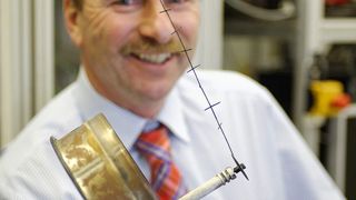 Dietmar Steiner von Bosch zeigt die von ihm erfundene Schnappelektrode. Wird der feine Draht unter Hochspannung gesetzt, holt er mehr als 85 % Feinstaub aus der Abluft von Kaminöfen. Die Elektrode ist an einer Metallscheibe befestigt, die sich beim Aufheizen und Abkühlen schlagartig verformt. So wird der gesammelte Staub wunschgemäß abgeschüttelt. Die große metallene Halterung hält die Elektrode im Abgasrohr. (Bild: Bosch)