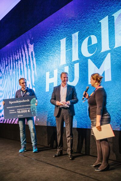 Stefan Hamann (l., Shopware) und Moderator Tobias Häusler überreichten Janin Bartoscheck von der Aktion Deutschland Hilft 10.000 Euro für ein Schulprojekt in Uganda. (Shopware AG)