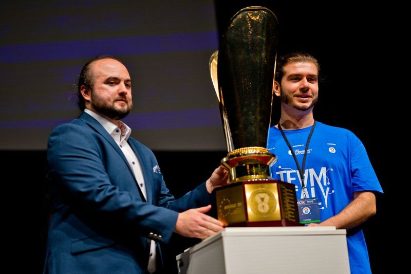 Der Captain des italienischen Teams (r.), das die ECSC 2025 in Warschau gewonnen hat, und Reich brachten zur Eröffnung der Challenge 2026 den Pokal auf die Bühne. (Bild: Bochum Wirtschaftsentwicklung | Donna & der Blitz)