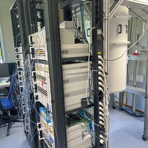 In der Realität sieht ein laufender Quantencomputer mit supraleitendem Quantenchip wenig aufregend aus, wie das Foto eines Systems von Peak Quantum in Garching bei München zeigt. Das komplexe Innenleben steckt in einer isolierten Hülle, die für die Kühlung auf eine Temperatur notwendig ist, die knapp über dem absoluten Nullpunkt liegt. Links daneben ist das Quantum Computing Control System von Zurich Instruments zu sehen, einem Tochterunternehmen von Rohde & Schwarz. (Bild: Vogel IT-Medien)