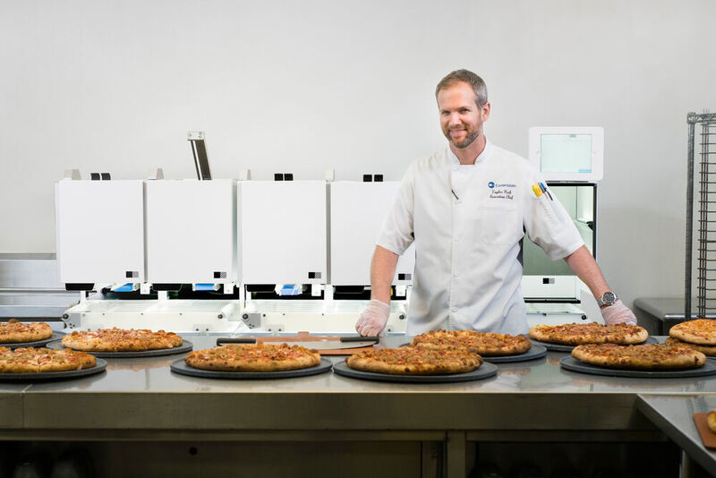 Picnic, ein Hersteller von Lebensmitteltechnologie mit Sitz in Seattle, gibt bekannt, dass sein automatisiertes Lebensmittelproduktionsystem vom Cateringunternehmen Centerplate ausgewählt wurde, um die Teilnehmer der CES 2020 mit Pizza zu verköstigen. Centerplate wird dazu den Picnic-Roboter, der bis zu 300 12-Zoll-Pizzen pro Stunde produzieren kann, auf der CES-Showfläche einsetzen. Das Bild zeigt Centerplate-CEO Taylor Park vor dem automatischen Pizza-Roboter von Picnic, den der Caterer im T-Mobile Park, der Heimat des Baseball-Teams Seattle Mariners, betreibt.  (Picnic)