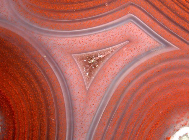 Achat: Fundort: Reichweiler bei Ida-Oberstein, Deutschland. Achat ist die gebänderte Varietät von Chalcedon, der wiederum eine mikrokristalline Varietät von Quarz darstellt. Der Grund für die rote Farbe bzw. Bänderung entpuppt sich unter dem Mikroskop als runde bis linsenförmige Aggregate von Hämatit. Die Natur des grünen, verzweigten Geflechts in der Bildmitte ist noch umstritten: während die herkömmliche Lehrmeinung darin anorganische Ausfällungen von gelöstem SiO2 vermuten, deuten neuere Untersuchungen eher auf eine organische Herkunft hin. Demzufolge wären diese Gebilde fossile Algen bzw. Bakterienkolonien, die bei Temperaturen von über 100 °C, die für die Bildung von Achat notwendig waren, leben konnten. Bildbreite: ca. 10 mm, Auflicht, Leica MZ16 Stereomikroskop (Michael Hügi, Schweizerisches Gemmologisches Institut)