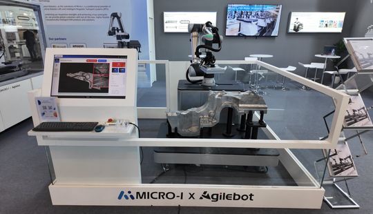 Ein Roboterarm führt hinter einer Schutzverglasung eine automatisierte Sichtprüfung an einem Metallbauteil durch, gesteuert über einen sichtbaren Prüfbildschirm.(Bild:  Micro-I)