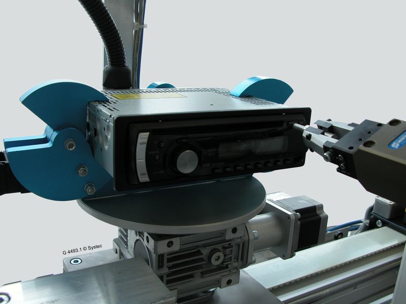 Ein elektrischer Greifer  nimmt im Systec-Messe-Exponat einen Metallstift auf und führt mit diesem verschiedene automatisierte Bedienaktionen am Autoradio aus, etwa das Betätigen des CD-Auswurf-Knopfes. (Archiv: Vogel Business Media)