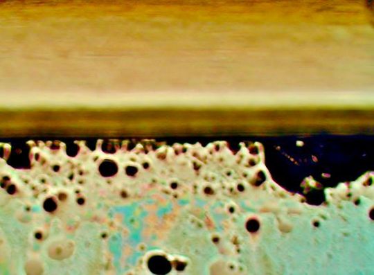 Das Bild zeigt in der Bildmitte die Laminat-Kante. Unterhalb ist der Blick durch den gebondeten Optical Stack mit sichtbar gemachtem Flüssig-Kleber und dessen Eigenschaften: schwarze Stellen zeigen massgeblich Blasenbildung (mehr ist schlechter), beige Stellen in unterschiedlichen Helligkeiten zeigen die Dichte-Verteilung des Klebers und Rot , Grün und Blau in Pastell-Tönen zeigen die Oberflächen-Benetzung und Haftkraft (mehr Farbe ist schlechter). (Bild:  Wammes und Partner)