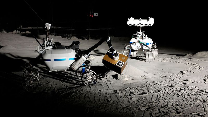Die beiden Rover LRU2 (links) und LRU1 sind in der LUNA-Halle unterwegs. LRU2 richtet mit seinem Roboterarm das LIBS (Laser-Induced Breakdown Spectroscopy) aus, das sich in der goldfarbenen Box befindet. LIBS verwendet einen gepulsten Laser, um zum Beispiel einen Stein zu analysieren. Forschende haben in der LUNA-Halle Messwerkzeuge und Roboter getestet, die Wassereis auf dem Mond finden können. (Bild: Roboter-Teamwork_ LRU1 und LRU2 arbeiten mit LIBS.jfif / DLR / CC BY 3.0 / dlr.de)