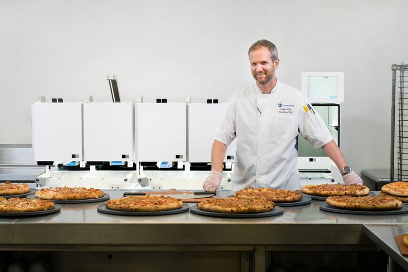 Picnic, ein Hersteller von Lebensmitteltechnologie mit Sitz in Seattle, gibt bekannt, dass sein automatisiertes Lebensmittelproduktionsystem vom Cateringunternehmen Centerplate ausgewählt wurde, um die Teilnehmer der CES 2020 mit Pizza zu verköstigen. Centerplate wird dazu den Picnic-Roboter, der bis zu 300 12-Zoll-Pizzen pro Stunde produzieren kann, auf der CES-Showfläche einsetzen. Das Bild zeigt Centerplate-CEO Taylor Park vor dem automatischen Pizza-Roboter von Picnic, den der Caterer im T-Mobile Park, der Heimat des Baseball-Teams Seattle Mariners, betreibt.  (Bild: Picnic)