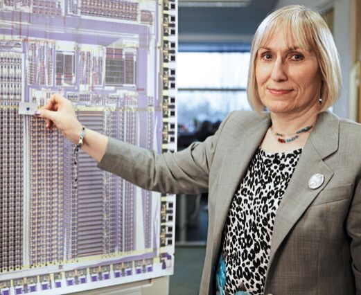 Sophie Mary Wilson hält 2013 vor ein vergrößertes Foto der ersten ARM-CPU ein Foto einer aktuellen ARM-Cortex-CPU im selben Maßstab. (Bild: Sophie Wilson / Chris Monk / CC BY-SA / Wikimedia Commons)
