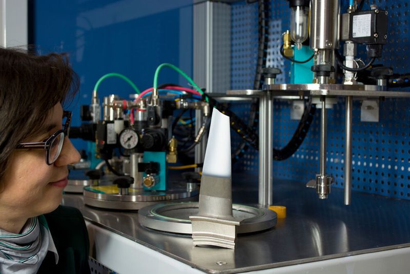 Dr. Laura Toma blickt auf eine mittels Suspensionsspritzen beschichtete Turbinenschaufel (im Hintergrund der am Fraunhofer IWS entwickelte Suspensionsförderer). Die Forscherin hat die Technologie des Suspensionsspritzens am Dresdner Institut etabliert und seit mehr als einem Jahrzehnt stetig weiterentwickelt. (Fraunhofer IWS)
