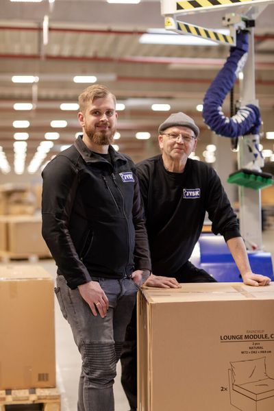 Rückenscherzen ausgemerzt! Von links: Bastian Böhm, Operations Manager bei JYSK, und Hans-Christian Hessler, dort Schwerbehindertenvertreter, im Homberger Logistiklager der JYSK Group. Rechst daneben der Vakuumschlauchheber Jumboflex Picker von Schmalz, der dort nun die Körperkräfte der Mitarbeiter schont. (Bild: Schmalz / M. Wagenhan)