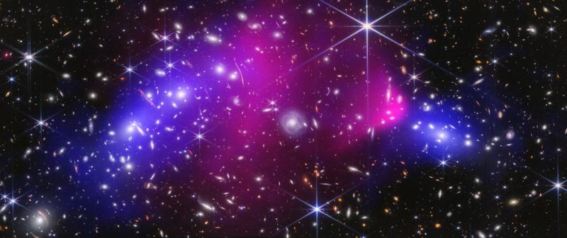 Rückschlag für Dunkle-Materie-Enthusiasten?  Der so genannte Bullet-Cluster galt bislang als Beweis für die Existenz Dunkler Materie. Vor rund vier Milliarden Jahren waren zwei Galaxiencluster mit mehr als 2.500 Kilometern pro Sekunde kollidierten, wobei sich das interstellare Gas von den Galaxienhaufen trennte. Bei den Galaxienhaufen beobachten Astronomen einen unerwartet starken Gravitationslinseneffekt, den sie bisher mit Dunkler Materie erklärten. Doch neue Daten stellen dies infrage. Ein internationales Forschungsteam unter Federführung der Universität Bonn hat aktuelle Aufnahmen des James-Webb-Weltraumteleskops analysiert und Argumente für eine alternative Theorie angeführt, die Modifizierte Newtonsche Dynamik (MOND), die ohne Dunkle Materie auskommt.  Demnach lasse sich der beobachtete Gravitationslinsen-Effekt durch die neu berechnete Menge von sichtbaren Sternen, Neutronensternen und Schwarzen Löchern erklären. Selbst, wenn man weiterhin an der Existenz Dunkler Materie festhalte, müsste man den neuen Daten zufolge deren postulierte Menge auf ungefähr die Hälfte reduzieren, betonen die Bonner Astronomen.  Eine Aufnahme des James-Webb-Weltraumteleskops oben zeigt die innere Region des Bullet-Clusters. Rosa: heißes Gas. Blau: postulierte Verteilung der Dunklen Materie.   Hier geht‘s zur Originalmeldung Wissenschaftsbild des Tages vom 9.7.2026  (Bild: NASA, ESA, CSA, STScI, CXC; Science: James Jee (Yonsei University, UC Davis), Sangjun Cha (Yonsei University), Kyle Finner (Caltech/IPAC))