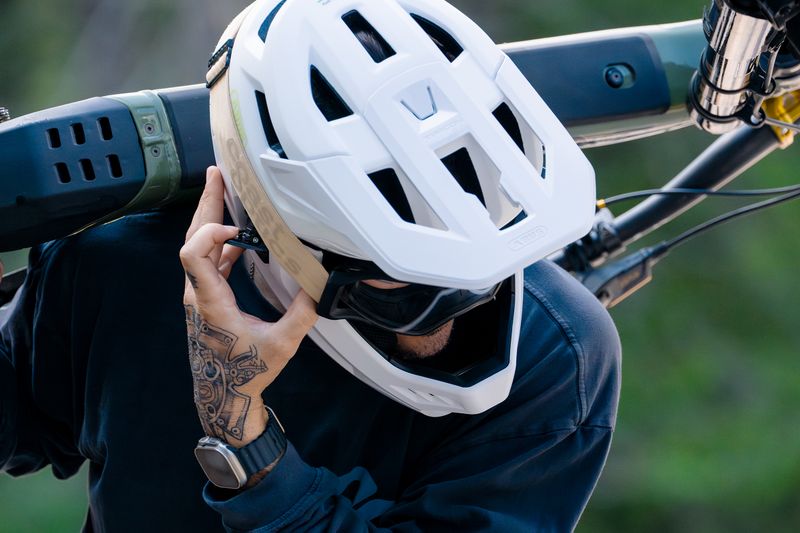 Mit dem „Targon“ stellt Abus einen modularen Fullface-Helm zum Mountainbiken vor. Sein besonderes Merkmal ist der abnehmbare Kinnbügel: Dank des einfach zu bedienenden Speed-Latch-Systems lässt sich der Schutz des Helmes flexibel an die jeweilige Strecke anpassen. So kann der Helm im Uphill als Halbschale und auf anspruchsvollen Trails, im Bikepark oder beim Downhilleinsatz als Fullface-Helm genutzt werden. Für einen festen und bequemen Sitz verwendet Abus ein Verstellsystem mit Drehrad am Hinterkopf – individuell verstellbare Wangenpolster sorgen dafür, dass die Fullface-Variante sicher und komfortabel sitzt. Die weichen Helmpolster verbessern den Tragekomfort und können bei Bedarf gewaschen oder ausgetauscht werden. Über den magnetischen Fidlock-Verschluss kann man den Helm mit einer Hand öffnen oder schließen. Unter dem Visier befindet sich zudem ein definierter Bereich zur sicheren Fixierung von Goggles, damit diese nicht verrutschen. Neben den gängigen Normen für Fahrradhelme erfüllt der Targon auch die Zusatznorm für Downhill-Helme und ist damit für den entsprechenden Einsatz freigegeben. Es gibt eine Version mit MIPS-Air-Node-Pro-Polster. Das MIPS-System ist darauf ausgelegt, bei einem schrägen Aufprall auf den Kopf wirkende Rotationskräfte zu reduzieren. Laut Hersteller liegt das Gewicht des Helms in der Größe S mit Kinnbügel bei 950 Gramm, ohne Kinnbügel bei 490 Gramm. Der Helm ist in den Farben „ash purple“, „lemon white“, „sand beige