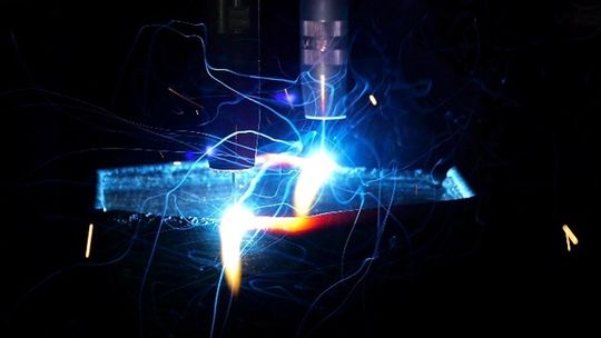 Beim drahtbasierten Additive Manufacturing (WAAM) erzeugt das Fronius iWave‑System hier einen intensiven Schweißlichtbogen und sichtbare Funken, während der Schweißbrenner Schicht für Schicht Metall auf das Substrat aufträgt, um schnell und materialeffizient ein Bauteil aufzubauen.(Bild:  ALOtec Dresden GmbH)