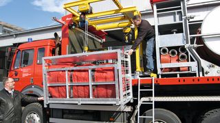 Zeigt einen Feuerwehr-LKW mit montiertem Halbportalkran der Feuerschutz Messer AG zum Be- und Entladen des LKWs mit Ladeplattformen mit Schlauch-Transportgestellen, Schlauchbrücken und Ausgleichsbehältern samt Zubehör. (Bild: Nozag)