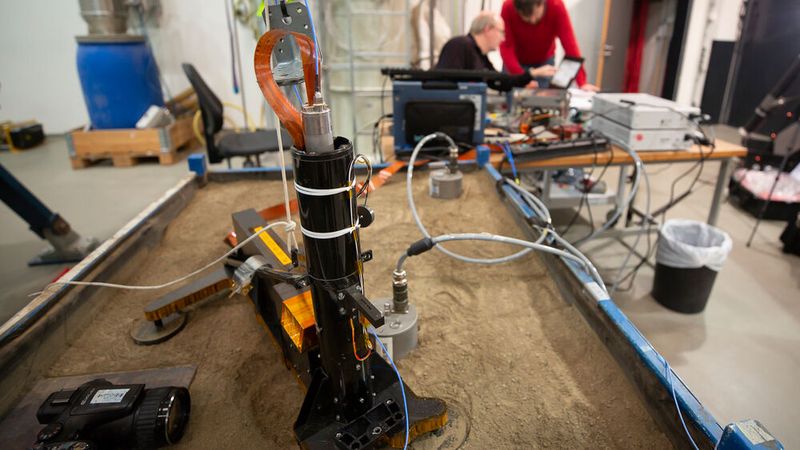 Das HP3-Experiment steht auf einer mit Sand gefüllten Box. Oben am Gehäuse ist ein Stück des Maulwurfs zu erkennen, der sich mit einem Schlagmechanismus allein in den Untergrund hämmern kann. (Bild: Testaufbau des HP3-Experiments am DLR in Bremen / CC BY 3.0)