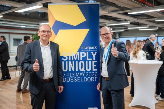 Interpack-Direktor Tomas Dohse (rechts) und Optima-CEO Dr. Stefan König (links) posieren vor dem Motto der Interpack: Simply Unique.(Bild:  Messe Düsseldorf)
