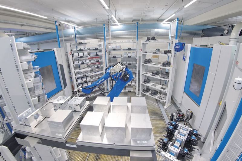 Blick in den Automationsbereich einer Fertigungsinsel mit zwei Heller «HF 3500». Gut zu erkennen sind die Paletten mit unterschiedlichen Roh- und Fertigteilen sowie der Knickarmroboter, der sowohl die Teile als auch die Paletten handeln kann. Im oberen Bild sind die beiden rückwärtigen Türen der Heller «HF 3500» zu erkennen, die über einen Schwenktisch verfügen und die Rohteile in den Bearbeitungsraum schwenken. Rechts unten sind darüber hinaus die verschiedenen Greifer für unterschiedliche Bauteile zu erkennen. (Bild: Matthias Böhm)