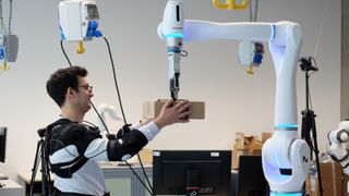 Forscher Federica Masiero nimmt ein Päckchen entgegen, dessen Gewicht der Roboter vorher bereits an das Exoskelett übermittelt hat. (Bild: Andreas Schmitz / TUM)