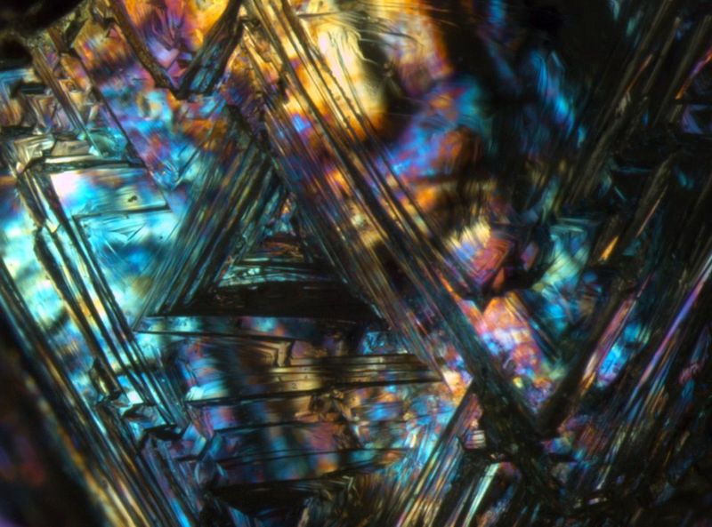 Natürliche Ätzstrukturen (so genannte Trigonen) auf Diamantkristall. Derartige Strukturen entstehen durch teilweise Auflösung des Diamants während des Aufstiegs aus dem oberen Erdmantel im heissen Kimberlitmagma. Die Interferenzfarben sind auf Spannungsdoppelbrechung zurückzuführen. Bildbreite: ungefähr 4 mm, Durchlicht, gekreuzte Polarisatoren, Leica MZ16 Stereomikroskop (Michael Hügi, Schweizerisches Gemmologisches Institut)
