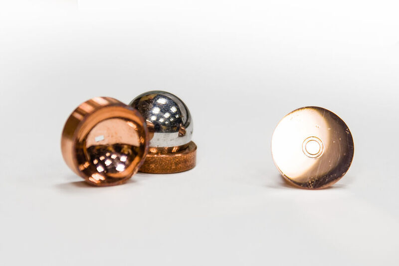 Copper mirrors direct and focus microwave photons in electrons on liquid helium system. The focusing mirror (on the left) is made by pressing a hard stainless steel ball (also shown in the picture) into a soft piece of copper. The flat mirror (on the right) shows two concentric circular electrodes (aka a Corbino pair) that are used to measure electron conductivity. (Olga Garnova / CC BY-SA 2.0)