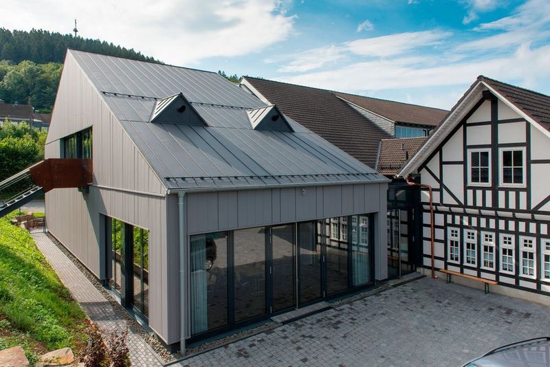 Das Dach des modernen Gebäudes ist in Falzanthrazit gehalten, es passt sich somit an die Schieferschindeln der Fachwerkhalle an. Die Fassade dagegen zeigt sich in hellerem Falzgrau. (Bild: Thyssenkrupp)