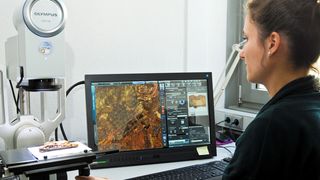 Abb. 2: Für die Restauratorin Ina Schneebauer-Meißner bedeutet die Arbeit mit modernster Mikroskop-Technik eine erhebliche Arbeitserleichterung. Auf dem Bildschirm im Hintergrund zeichnet sich ein Webmuster ab – Überreste von fast 1.500 Jahre alten Textilfasern. (Bild: L. Ruminski, Bremen)