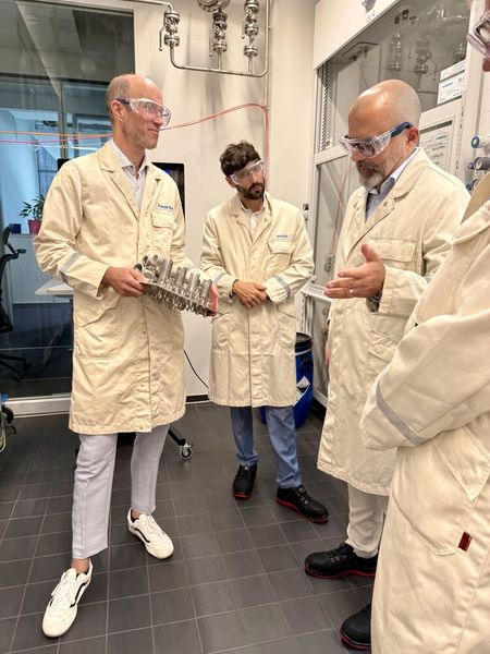 Sebastian Härtner (MTP-Experte bei Merck, links) zeigt dem hessischen Staatsekretär Daniel Köfer (rechts) und dem Landtagsabgeordneten Bijan Kaffenberger (Mitte) ein Reaktionsmodul bei einem Besuch im August 2026. (Bild: Merck)
