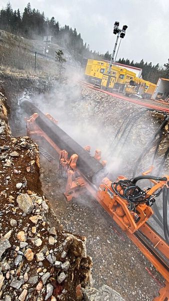 Die Druckluft aus den Atlas-Copco-Kompressoren (im Hintergrund) treibt den Schlagkolben des Imlochhammers im Inneren des Hammerbohrgeräts (groß im Bild) an, wodurch der Bohrkopf der Maschine auf die Bohrlochsohle schlägt. Durch die Schläge tritt am Kopf Luft aus, die zugleich die Sohle reinigt und das gelöste Bohrgut über den Ringraum der Maschine nach hinten und zurück an die Erdoberfläche spült.  (Bild: Petzold)