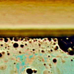 Das Bild zeigt in der Bildmitte die Laminat-Kante. Unterhalb ist der Blick durch den gebondeten Optical Stack mit sichtbar gemachtem Flüssig-Kleber und dessen Eigenschaften: schwarze Stellen zeigen massgeblich Blasenbildung (mehr ist schlechter), beige Stellen in unterschiedlichen Helligkeiten zeigen die Dichte-Verteilung des Klebers und Rot , Grün und Blau in Pastell-Tönen zeigen die Oberflächen-Benetzung und Haftkraft (mehr Farbe ist schlechter). (Bild:  Wammes und Partner)