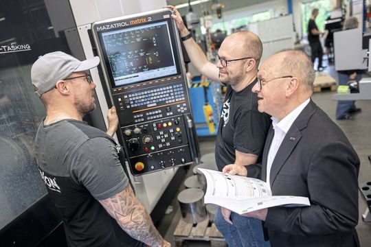 Thomas Lomoth (PPA, Metaxon), David Willing (Geschäftsführer, ZE Zerspanungsexperten GmbH, Walter-Vertriebspartner) und Josef Schmidt (Technische Beratung, Walter AG) diskutieren den neuen Dreh-Fräsprozess vor dem Mazak-Multitaskzentrum. Unten mittig im Bild die Rohteile aus dem Rostfrei-Stahl.(Bild:  Walter AG)