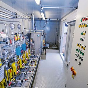 Innenansicht einer Ammoniak-Dosierstation mit zehn Kolbenmembran-Pumpen. Ganz hinten befindet sich eine Aufstellfläche für den IBC. Von dort wird die Ammoniaklösung per Deionatstrom in den Ansetzbehälter (Mitte) gesaugt und dabei verdünnt. IBC-Gestell, Ansetzbehälter und Dosierpumpen befinden sich in einer gemeinsamen Auffangwanne mit Leckagesensor.