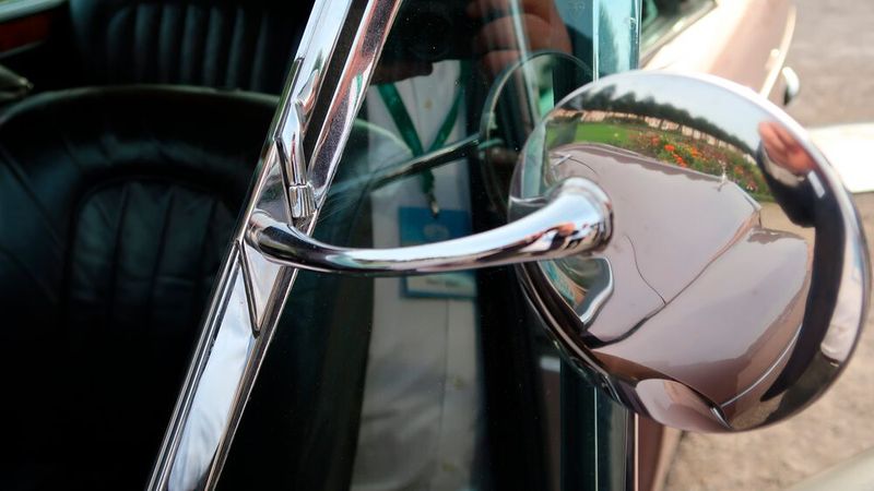 In früheren Jahren wurden Außenrückspiegel meist per Bohrmaschine nachgerüstet. Heute ist man vorsichtiger und vermeidet Bohrlöcher, die beim Rückbau als Verluststellen in der Karosserie verbleiben. Hier erfolgte die Nachrüstung besonders geschickt: Der Spiegel wurde an einer dünnen Metallschiene befestigt, die wiederum am vorderen Türrahmen und – im Bild zu sehen – am oberen Drehpunkt des Dreieckfensters fixiert ist. Durch die Verchromung wirkt alles als ein Teil des Fensterrahmens. Gesehen an einem Rolls-Royce Silver Cloud III „Chinese Eyes“ Mulliner Coupé aus dem Jahr 1965. (Bild: Peter Diehl)
