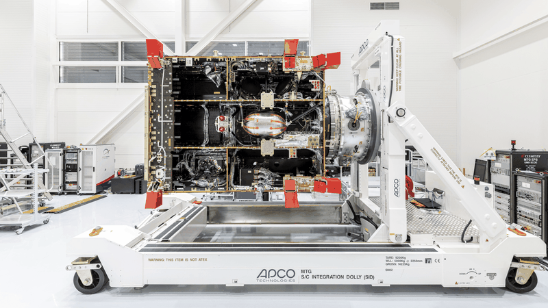 A large MTG-I satellite platform/bus in a cleanroom, mounted horizontally on an APCO Technologies “MTG S/C Integration Dolly (SID)”. The spacecraft’s internal structure, harnessing, and thermal insulation are visible, with red protective covers on multiple interfaces.(Source:  OHB SE)