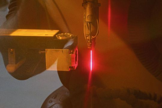 Die Qualität der Düsenöffnung wird indirekt durch Analyse der Raman-Strahlung erfasst (das ist gestreutes Laserlicht, dessen Frequenzunterschied zum ursprünglichen Strahl zur Analyse dient). Links der Sensor, rechts der durch das Laserschutzfenster rot erscheinende LMJ-Strahl. (Bild:  Synova)