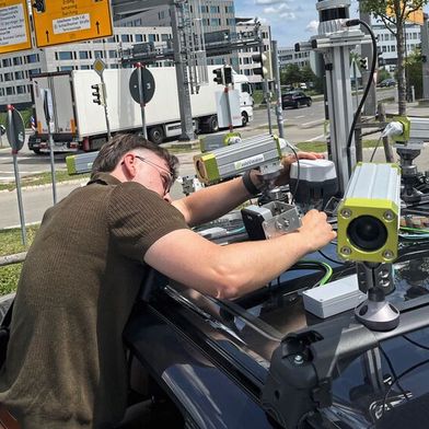 TUM-Forscher Erik Leo Haß prüft die Sensorik auf dem Fahrzeug an einer Kreuzung in Garching-Hochbrück. Aus den unterschiedlichen Echtzeit-Messungen von Kameras, Lidaren und Radaren wird ein permanent aktualisiertes Bild der Verkehrssituation erstellt. (Bild: A.Schmitz / TUM)