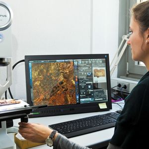 Abb. 2: Für die Restauratorin Ina Schneebauer-Meißner bedeutet die Arbeit mit modernster Mikroskop-Technik eine erhebliche Arbeitserleichterung. Auf dem Bildschirm im Hintergrund zeichnet sich ein Webmuster ab – Überreste von fast 1.500 Jahre alten Textilfasern.