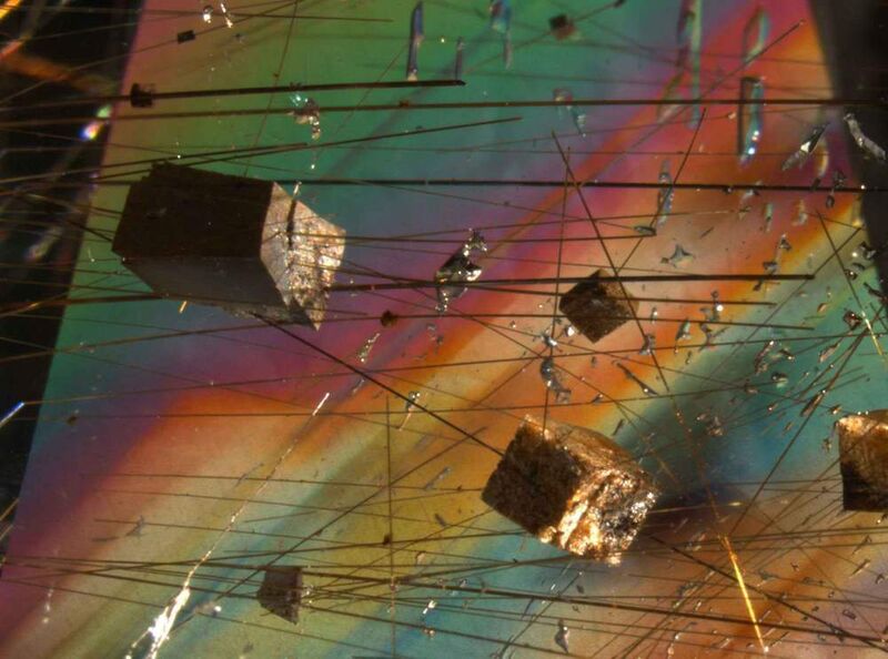 Einschlüsse von vermutlich Siderit (FeCO3) und Rutilnadeln in Bergkristall (Quarz). Fundort: Minas Gerais, Brasilien. Bei den braunen, rhomboedrischen Kristallen handelt es sich aufgrund der Morphologie und Farbe höchstwahrscheinlich um das Eisencarbonat Siderit. Die starken Interferenzfarben des ansonsten farblosen Quarzes entstehen durch die fast parallele Blickrichtung zur optischen Achse des Kristalls im polarisierten Licht. (Bildhöhe: ca. 10 mm; Kombination von Auflicht (Glasfaserlicht) und Durchlicht; gekreuzte Polarisatoren; Bild: Michael Hügi, Schweizerische Gemmologische Gesellschaft SGG) (Archiv: Vogel Business Media)