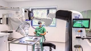 Dr. Maximilian Kückelhaus präsentiert bei einem „Trockentraining“ das neue Verfahren. Der Operationsroboter (links) ist mit einem robotischen Mikroskop (rechts) vernetzt.  (Bild: WWU - Peter Leßmann)