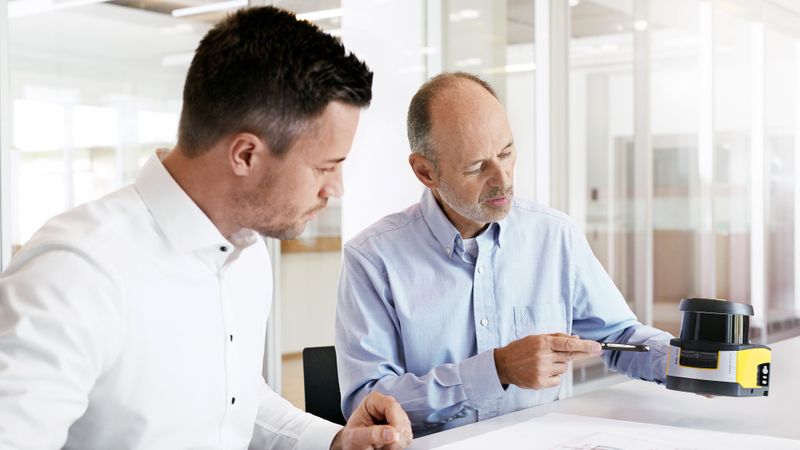 Im Austausch über Maschinensicherheit: Zwei Experten prüfen einen Sicherheitssensor und technische Planungsunterlagen – ein wichtiger Schritt für sichere Maschinen und die Umsetzung neuer Anforderungen.(Bild:  Leuze Electronic GmbH + Co. KG)