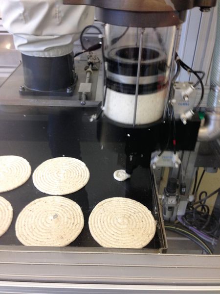 Mit einer Dosiereinheit dressiert der Roboter in kreisenden Bewegungen die Haselnuss-Baisermasse. Das volle Backblech schiebt ein Mitarbeiter in den Ofen und ein leeres wird von einem Stapel automatisch in die Roboterzelle gezogen. (Bild: Suteria)