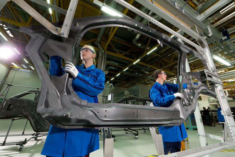 Zwei BMW-Mitarbeiter bereiten einen CFK-Seitenrahmen für die mechanische Bearbeitung im Wasserstrahlschneidverfahren vor. (Bild: BMW)