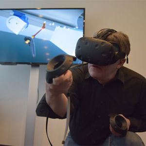 Ein Trainingsteilnehmer hat die VR-Brille auf und führt im Training mit den Controllern die Wartungsarbeiten aus. Im Hintergrund dieses Fotos ist auf dem LCD-Monitor zu sehen, was er gerade über die Brille wahrnimmt.