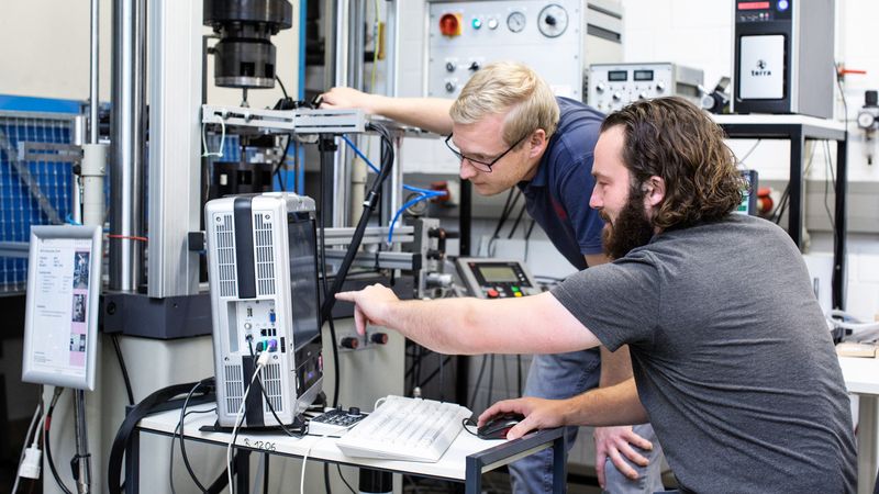 Dr.-Ing. Philipp Krooß und Dr.-Ing. Christian Lauhoff (von links) am servohydraulischen Prüfstand des Fachgebiets. Dieser Versuchsaufbau war eine zentrale Säule für die Studie: Er ermöglicht es, die mechanischen Eigenschaften des Materials zu untersuchen und gleichzeitig die Veränderungen im Gefüge unter dem Lichtmikroskop zu beobachten.(Bild:  Christine Buhl/Uni Kassel)