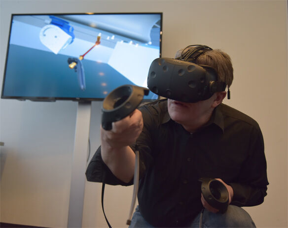 Ein Trainingsteilnehmer hat die VR-Brille auf und führt im Training mit den Controllern die Wartungsarbeiten aus. Im Hintergrund dieses Fotos ist auf dem LCD-Monitor zu sehen, was er gerade über die Brille wahrnimmt. (Tema)