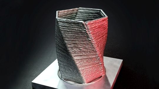 Das additiv gefertigte Musterbauteil zeigt den schichtweisen WAAM‑Aufbau: ein sechseckiger, leicht verdrehter Metallbecher mit deutlich sichtbaren, übereinanderliegenden Schweißraupen, auf einer Trägerplatte gefertigt.(Bild:  ALOtec Dresden GmbH)