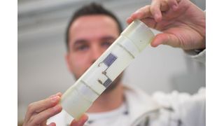 Michael Heinrich von Fiber Check zeigt den gestickten Dehnungssensor, für den ein Metallfaden in ein Vlies und dann in ein Glasfaserbauteil eingearbeitet wird - dieses System erkennt Schäden in Rotorblättern schon bei der Entstehung.  (Bild: TU Chemnitz/Heiko Kießling)